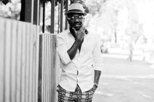 Stylish african american man in white shirt and colored pants with hat and glasses posed outdoor. Black fashionable model boy. photo