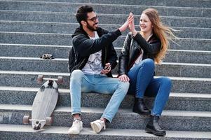 Cool multiracial couple sitting on stairs with longboard and give high five each other. photo