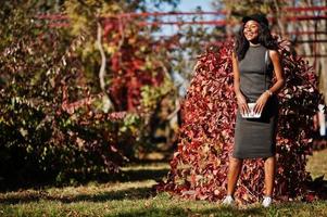 Stylish african american girl in gray tunic, crossbody bag and cap posed at sunny autumn day against red leaves. Africa model woman. photo