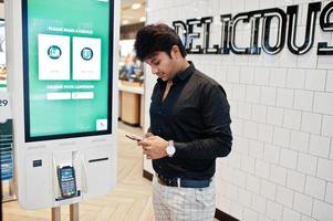 Indian man customer at store place orders and pay through self pay floor kiosk for fast food, payment terminal. His looking at mobile phone. Make a choise of language on screen. photo