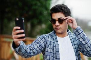 Stylish indian young man at sunglasses wear casual posed outdoor and making selfie at mobile phone. photo