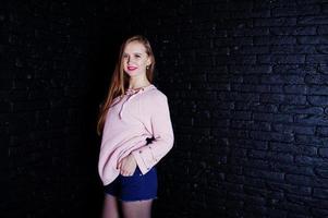Studio shot of brunette girl at pink sweater with jeans shorts against black brick wall. photo