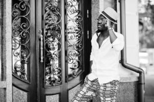 Stylish african american man in white shirt and colored pants with hat and glasses posed outdoor. Black fashionable model boy. photo