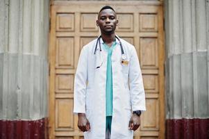 médico afroamericano masculino en bata de laboratorio con estetoscopio al aire libre contra la puerta de la clínica. foto