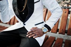 Chic handsome african american man in white suit sitting on bench. Close up photo of golden watches on hand.