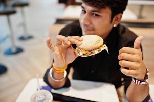Stylish indian man sitting at fast food cafe and eating hamburger. Show thumb up. photo
