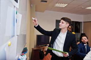 Mixed race business coach presenting report standing near whiteboard pointing on sales statistic shown on diagram and chart teach diverse company members gathered together in conference room. photo