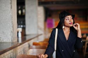 encantadora modelo afroamericana con chaqueta negra, sombrero y riñonera relajándose en el café durante el tiempo libre. foto