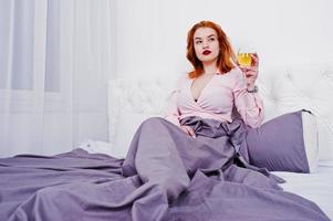 Hermosa chica pelirroja con blusa rosa y falda roja con una copa de vino a mano en la cama de la habitación. foto