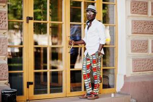 Stylish african american man in white shirt and colored pants with hat and glasses posed outdoor. Black fashionable model boy. photo