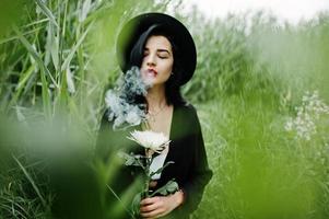 Sensual smoker girl all in black, red lips and hat. Goth dramatic woman hold white chrysanthemum flower and smoking on common reed. photo