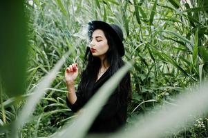 Sensual smoker girl all in black, red lips and hat. Goth dramatic woman smoking on common reed. photo