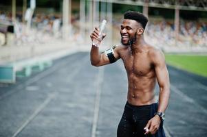 African american male athlete sexy sport bare torso man with running sports arm case for mobile phone, posed at stadium and drinking water from bottle. photo