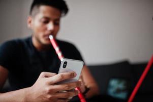 Handsome and fashionable indian man in black sitting at room, smoking hookah and looking on mobile phone. photo