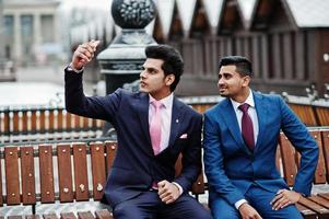 Two elegant indian fashionable mans model on suit posed at winter day sitting on bench and making selfie. photo