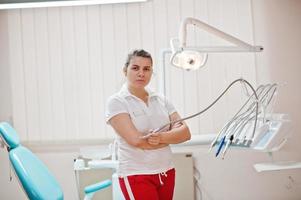 Portrait of female dentist woman crossed arms standing in her dentistry office near chair. photo