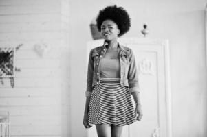 Attractive african american woman with afro hair wear on skirt and jeans jacket, posed at white room. Fashionable black model. photo