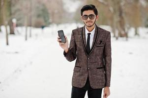 Stylish indian student man in brown suit and sunglasses posed at winter day outdoor with mobile phone at hand. photo