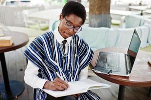 African man in traditional clothes and glasses sitting behind laptop at outdoor caffe and writing something on his notebook. photo