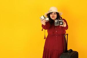 atractiva mujer viajera del sur de asia con vestido rojo intenso, sombrero posado en el estudio sobre fondo amarillo con maleta y cámara de fotos antigua.