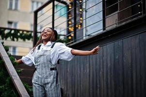 mujer afroamericana en overoles y boina posada en la terraza al aire libre con guirnaldas de adornos navideños. foto