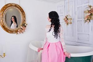 Young brunette girl in pink skirt and white blouse posed indoor against room with decor bath and mirror. photo