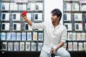 Indian man customer buyer at mobile phone store with wireless earphones and speaker sitting on chair. South asian peoples and technologies concept. Cellphone shop. photo