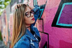chica hipster casual con estilo en ropa de jeans y gafas contra una gran pared de graffiti. foto