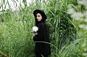Sensual girl all in black, red lips and hat. Goth dramatic woman on common reed hold white chrysanthemum flower. photo