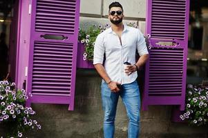 Elegante modelo de hombre árabe alto con camisa blanca, jeans y gafas de sol en la calle de la ciudad. Barba atractivo chico árabe con una taza de café contra ventanas violetas. foto