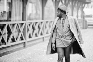 elegante modelo afroamericano con abrigo gris, corbata de chaqueta y sombrero rojo posado en la calle con niebla. foto en blanco y negro.