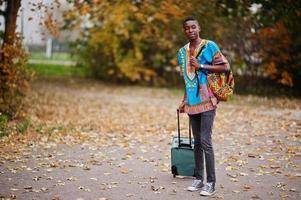 African man in africa traditional shirt on autumn park with backpack and suitcase. Emigrant traveler. photo