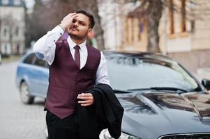 Elegante hombre de negocios indio con traje formal de chaleco de pie contra un coche de negocios negro en la calle de la ciudad. foto