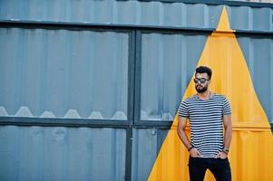 Handsome tall arabian beard man model at stripped shirt posed outdoor. Fashionable arab guy at sunglasses against yellow painted wall. photo