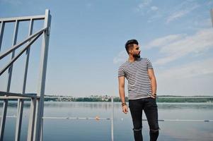 Handsome tall arabian beard man model at stripped shirt posed outdoor on pier of lake. Fashionable arab guy. photo