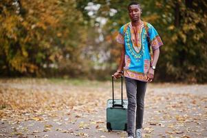 hombre africano en camisa tradicional africana en el parque de otoño con mochila y maleta. viajero emigrante. foto