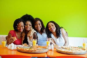 Four young african girls in bright colored restaurant eating pizza and having fun together. photo