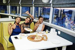 tres mujeres africanas vestidas posando en el restaurante, comiendo pizza y bebiendo jugo. foto