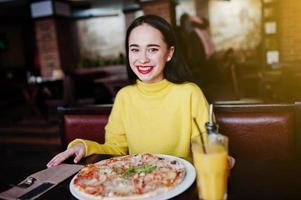 chica morena divertida en suéter amarillo comiendo pizza en el restaurante. foto