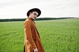 hombre elegante con gafas, chaqueta marrón y sombrero posado en campo verde. foto