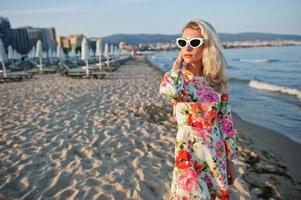 Blonde woman wearing dress and sunglasses standing on sand beach at sea shore enjoying view of sunset. photo