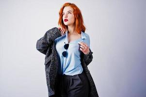 Portrait of a fantastic redheaded girl in blue shirt, grey overcoat posing with sunglasses in the studio. photo