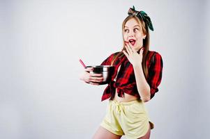 joven ama de casa divertida en camisa a cuadros y pantalones cortos amarillos pin up estilo con cacerola y cuchara de cocina aislado sobre fondo blanco. foto