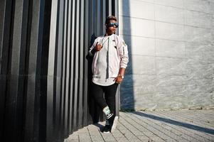 hombre árabe con estilo hipster en gafas de sol posó al aire libre en la calle al atardecer contra la pared gris. foto