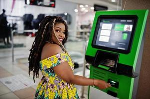 linda chica afroamericana de pequeña altura con rastas, vestida con un vestido amarillo de color, contra un cajero automático con tarjeta de crédito a mano. foto