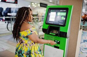 Cute small height african american girl with dreadlocks, wear at coloured yellow dress, against ATM with credit card at hand. photo