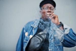 African american man in jeans jacket, beret and eyeglasses, smoking cigar on gray steel background. photo