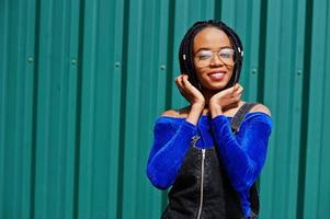 African woman in denim overalls skirt, eyeglasses posed against green steel wall. photo