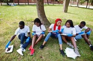 grupo de felices voluntarios africanos sentados bajo un árbol en el parque y escribiendo algo en portapapeles. Concepto de voluntariado, caridad, personas y ecología de África. foto