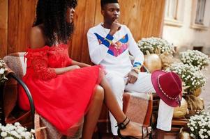 elegante pareja afro francia de moda posaron juntos en el día de otoño. modelos africanos negros enamorados sentados contra una decoración de madera con flores y calabazas. foto